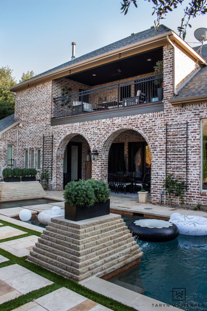 Tour this Modern English Country Backyard featuring classic black and white outdoor furniture and decor with greenery and brick exterior, oval shaped pool with pavers and turf. 