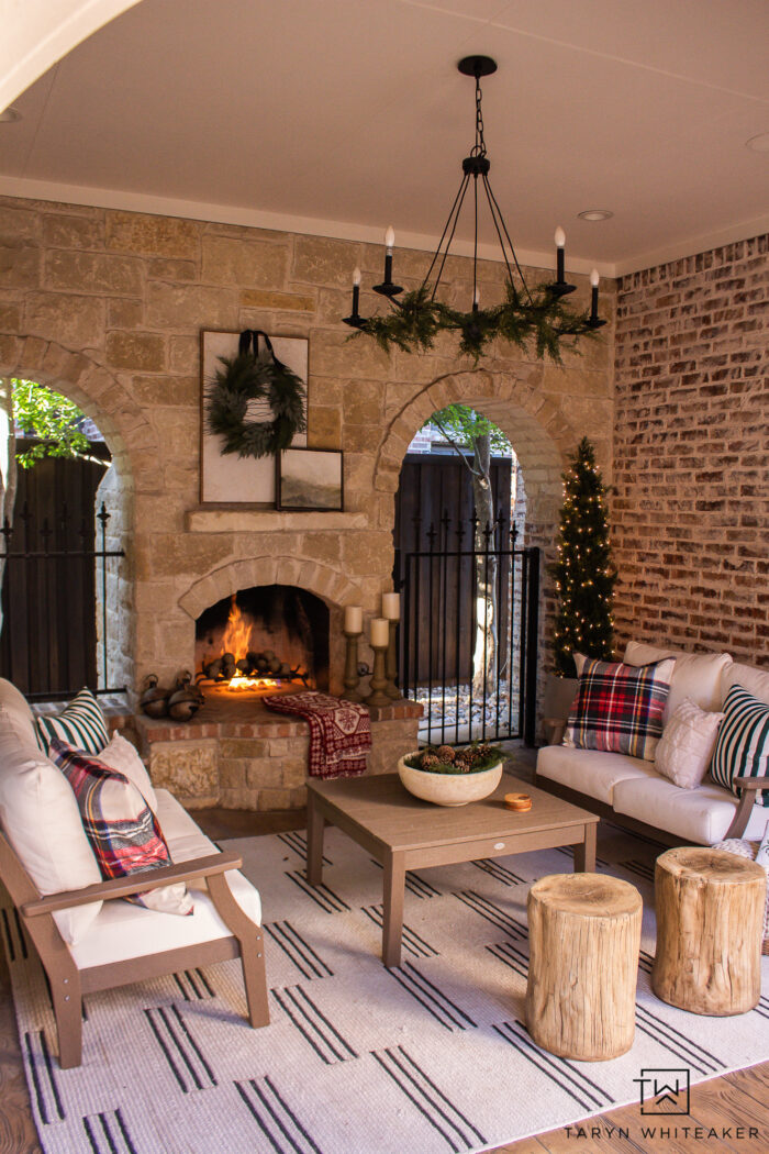 Outdoor Fireplace Seating area decorated for the holidays with pops of tartan mixed green velvet pillows and modern stripes, paired with faux cedar trees wrapped in lights.