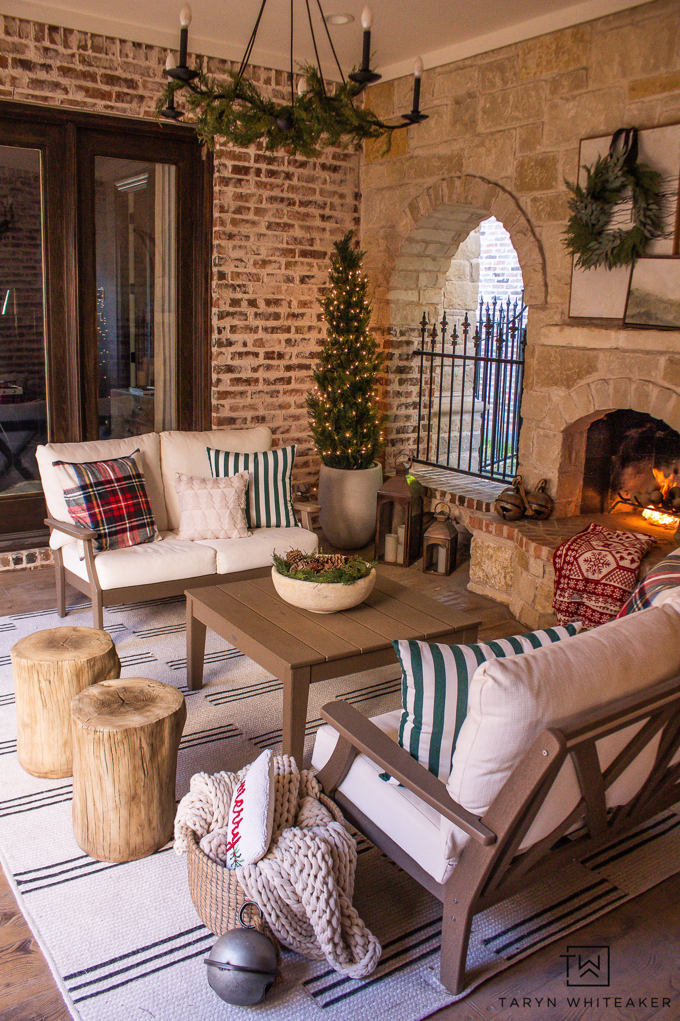 Outdoor Fireplace Seating area decorated for the holidays with pops of tartan mixed green velvet pillows and modern stripes, paired with faux cedar trees wrapped in lights.